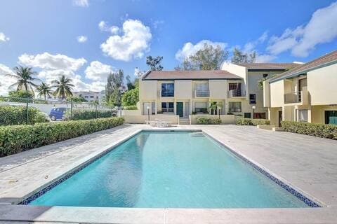 The outdoor area features a spacious swimming pool surrounded by a concrete deck. Lush greenery and palm trees provide a tropical backdrop, while a two-story condominium building is situated behind the pool, reflecting a calm and inviting atmosphere.