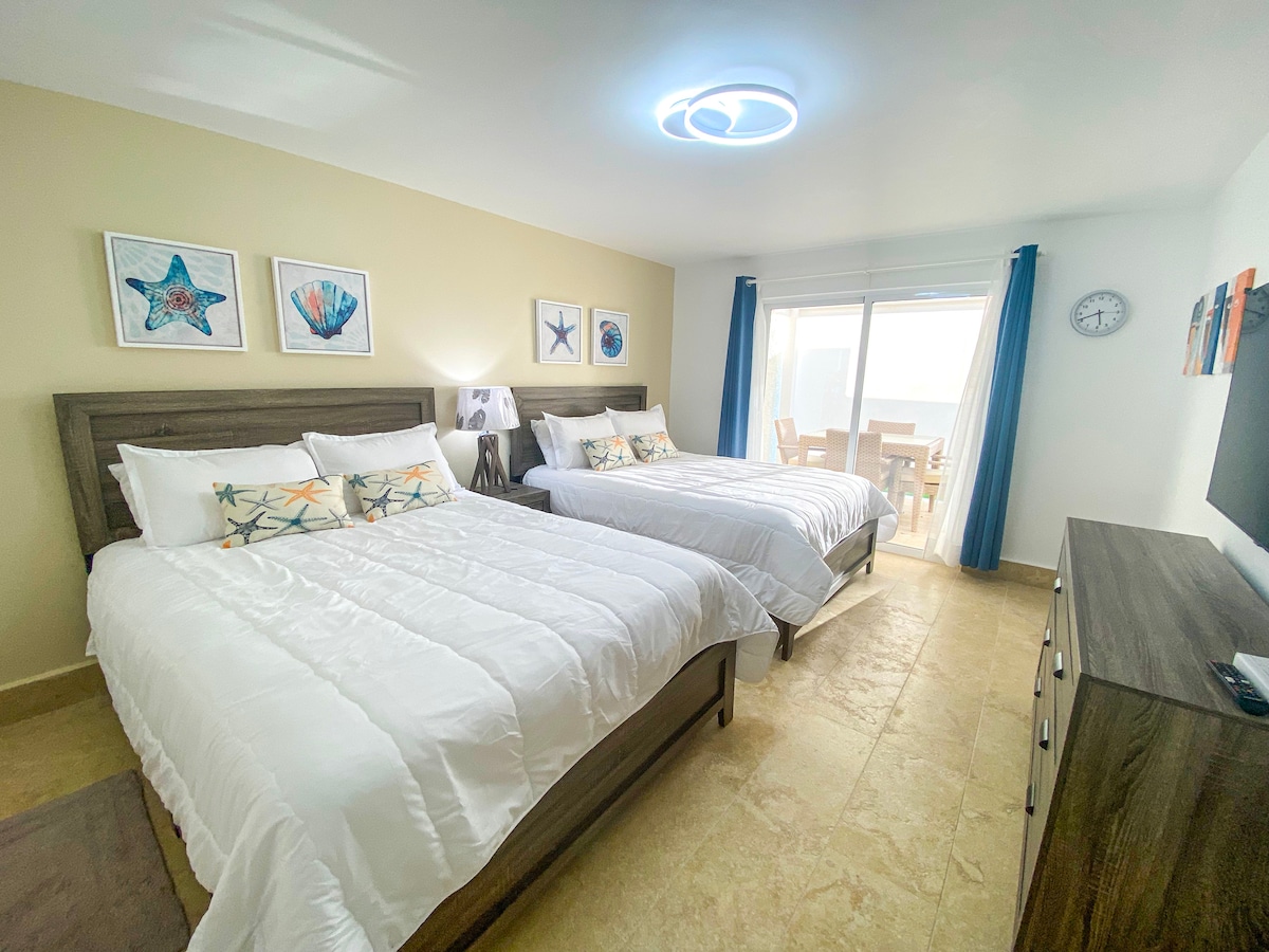 A spacious bedroom features two large beds with crisp white bedding, complemented by decorative pillows. Artwork depicting sea life adorns the walls. Natural light filters in through large sliding doors that lead to a private outdoor area, while a flat-screen TV is mounted on the opposite wall.
