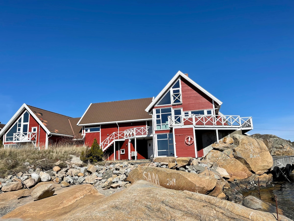 The exterior of a red wooden building is shown, featuring a contemporary design with large windows and a sloping roof. The structure is set against a clear blue sky and surrounded by rocky terrain, with a wooden walkway leading to the entrance.