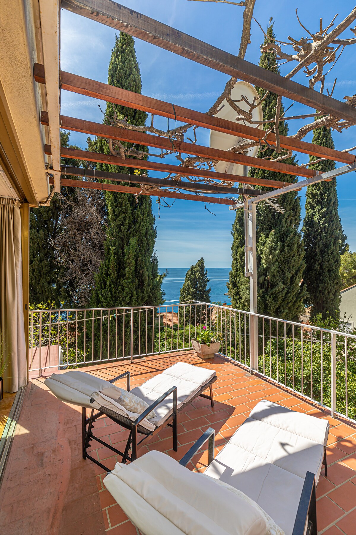 A terrace offers two lounge chairs under a wooden pergola, with a view of the sea. Lush greenery is visible in the background, and terracotta tiles provide a warm base. Sunlight brightens the space, creating an inviting atmosphere.