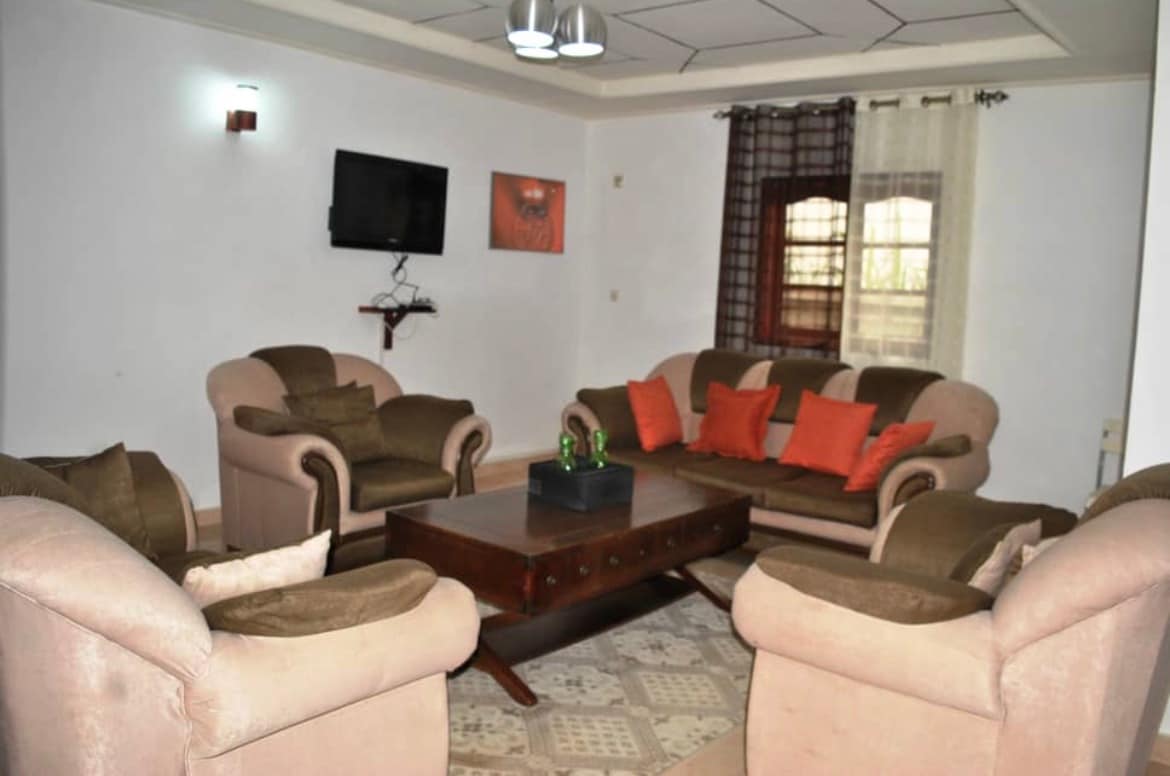A cozy living room features a comfortable seating arrangement with several sofas and armchairs, accented with orange cushions. A wooden coffee table sits in the center, and a mounted television is visible on the wall. Natural light enters through a window adorned with sheer curtains.