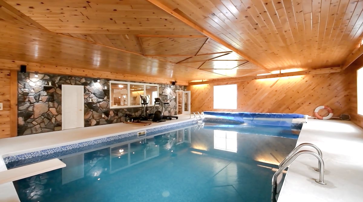 An indoor heated saltwater pool is featured, surrounded by wooden paneling and stone accents. The pool measures 3-8 feet deep and features a smooth, tiled edge. Large windows allow natural light to illuminate the space, creating a serene atmosphere for relaxation.