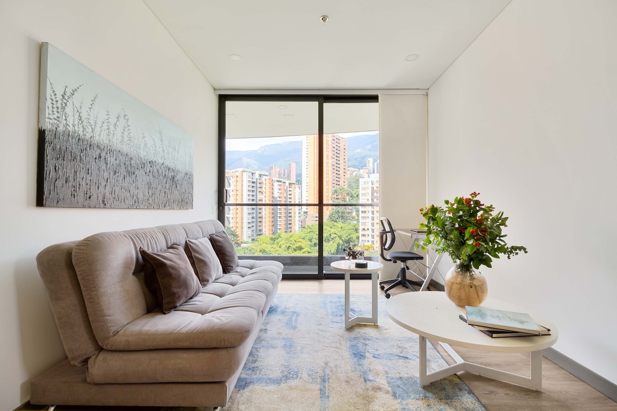 A modern living space features a comfortable gray sofa with decorative pillows. A round coffee table is set beside a floral arrangement, while a desk and chair are positioned nearby. Large windows reveal a city view with surrounding greenery.