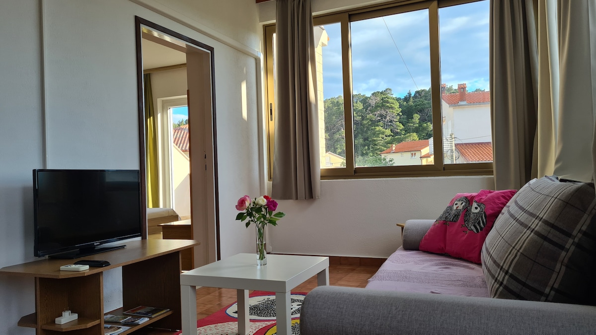 A living room is highlighted by large windows, allowing natural light to fill the space. A cozy sofa is positioned beside a small coffee table, while a television is placed on a wooden stand. Decorative flowers in a vase add a touch of color to the room.