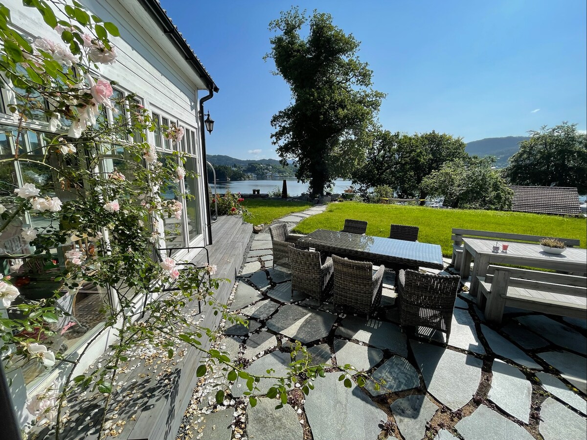 A spacious outdoor patio features a large dining table surrounded by comfortable wicker chairs. Natural stone flooring complements the lush green lawn and rose bushes, with views of the nearby sea and trees creating a peaceful setting.