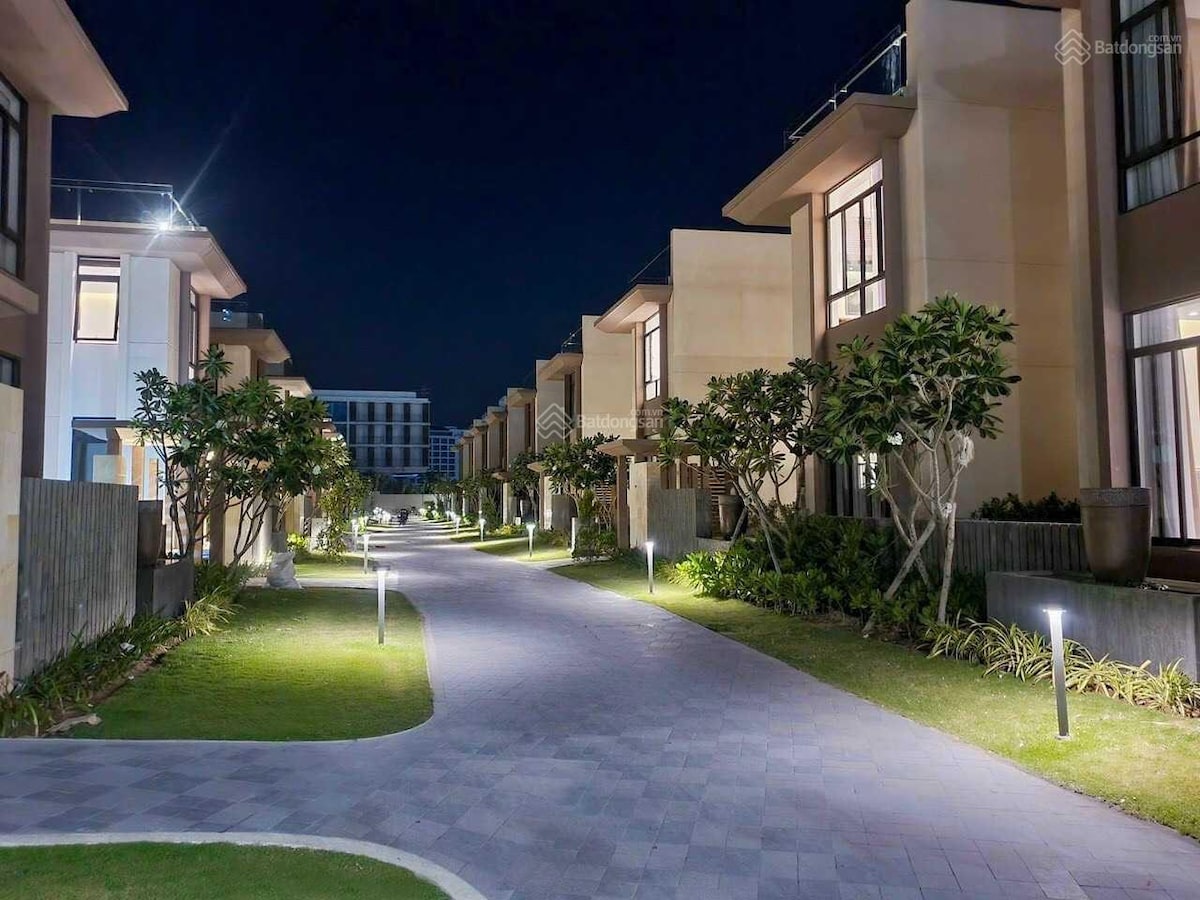 A well-lit pathway is framed by neatly arranged trees and landscaped gardens. The modern buildings are situated on either side, showcasing large windows that reflect the soft glow of the outdoor lighting. A serene atmosphere is created for evening strolls.