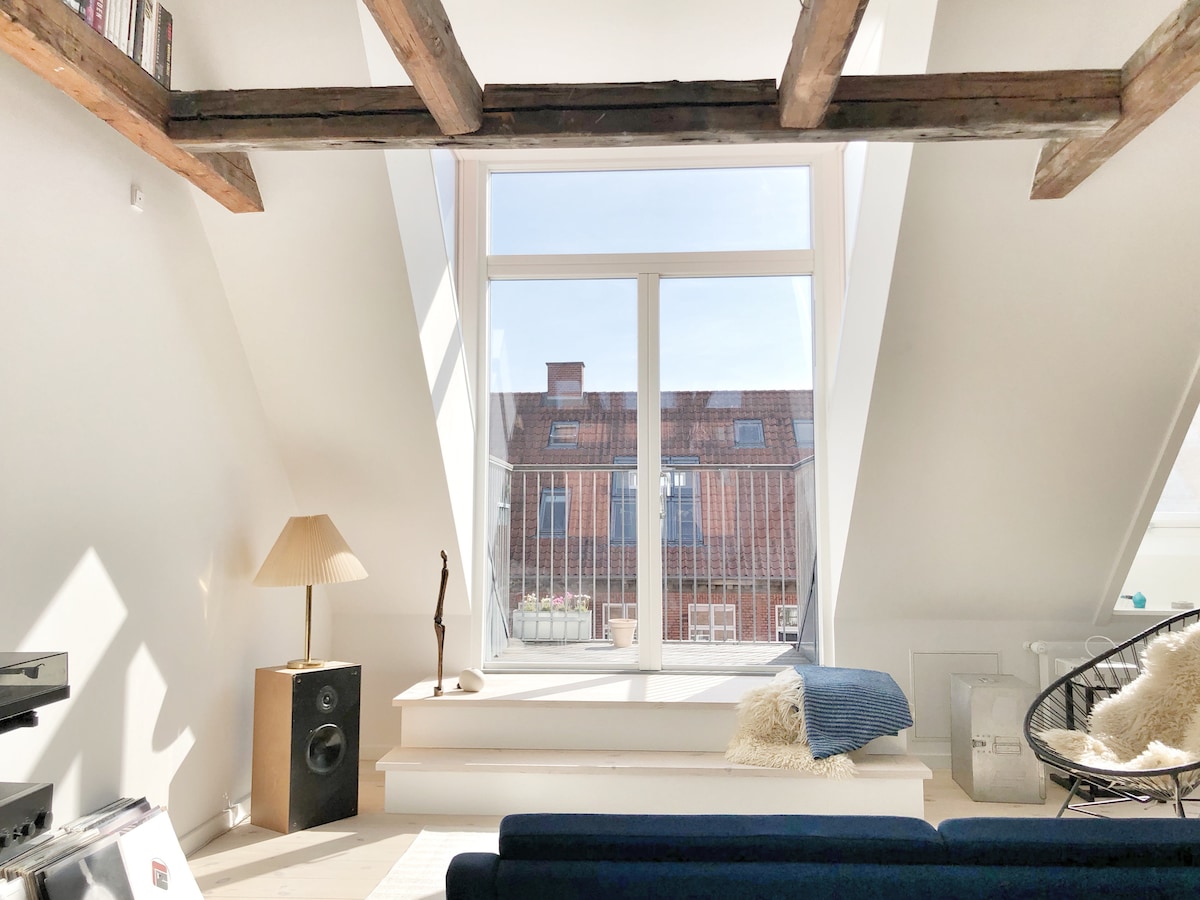 A bright room features multiple windows that allow ample natural light, highlighting the sloped ceiling with exposed wooden beams. A comfortable seating area is situated near the window, complemented by a tall lamp and decorative items, creating an inviting space for relaxation.