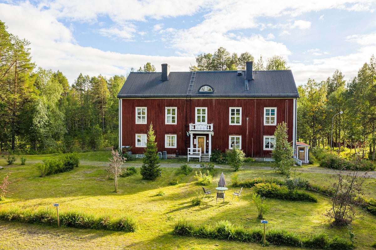 A spacious, two-story red wooden house is surrounded by landscaped greenery. Large windows and a central dormer enhance its structure. The front yard features a seating area with chairs positioned near a fire pit, inviting a connection with the natural surroundings.