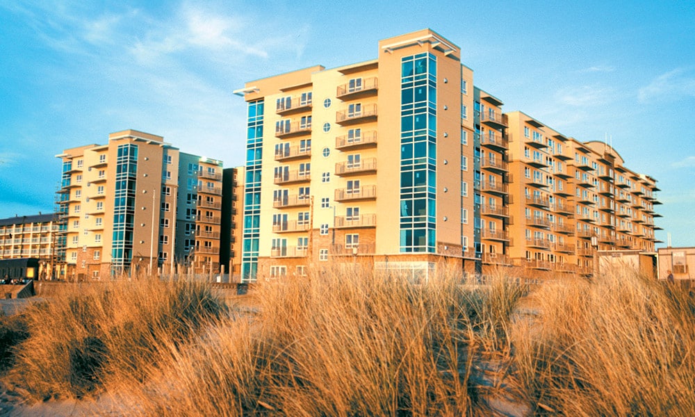 A modern beachfront resort is viewed from the sandy beach, featuring multiple stories with glass balconies that reflect the sunlight. The building's warm tones complement the natural landscape of tall grasses in the foreground, contributing to the coastal scenery.