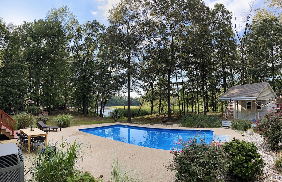 An outdoor area features a salt-water pool surrounded by lush greenery, with trees providing natural shade. A small cabin is visible to the right, and the river can be seen in the background. Comfortable seating options are situated near the poolside.