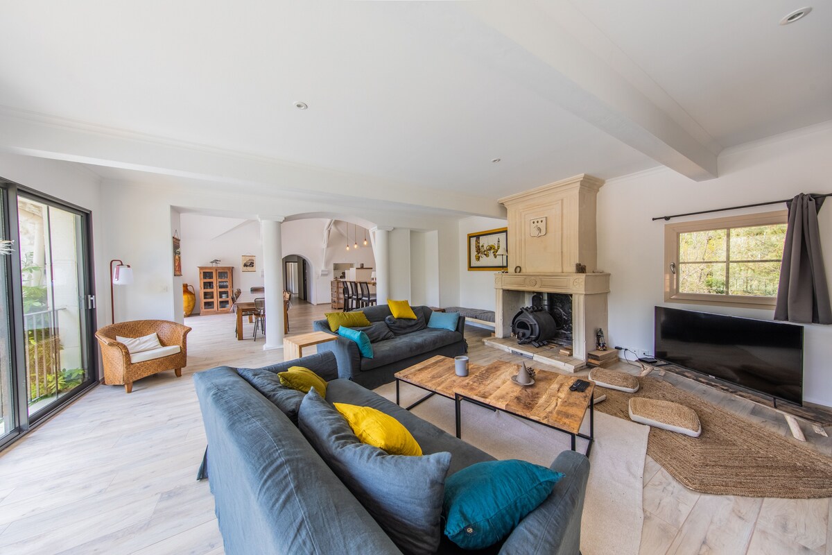 A spacious living area features comfortable seating with blue couches and vibrant yellow cushions. Natural light floods the room through large glass doors, revealing a cozy fireplace and a wooden coffee table. A large television is positioned against one wall, creating a welcoming space.