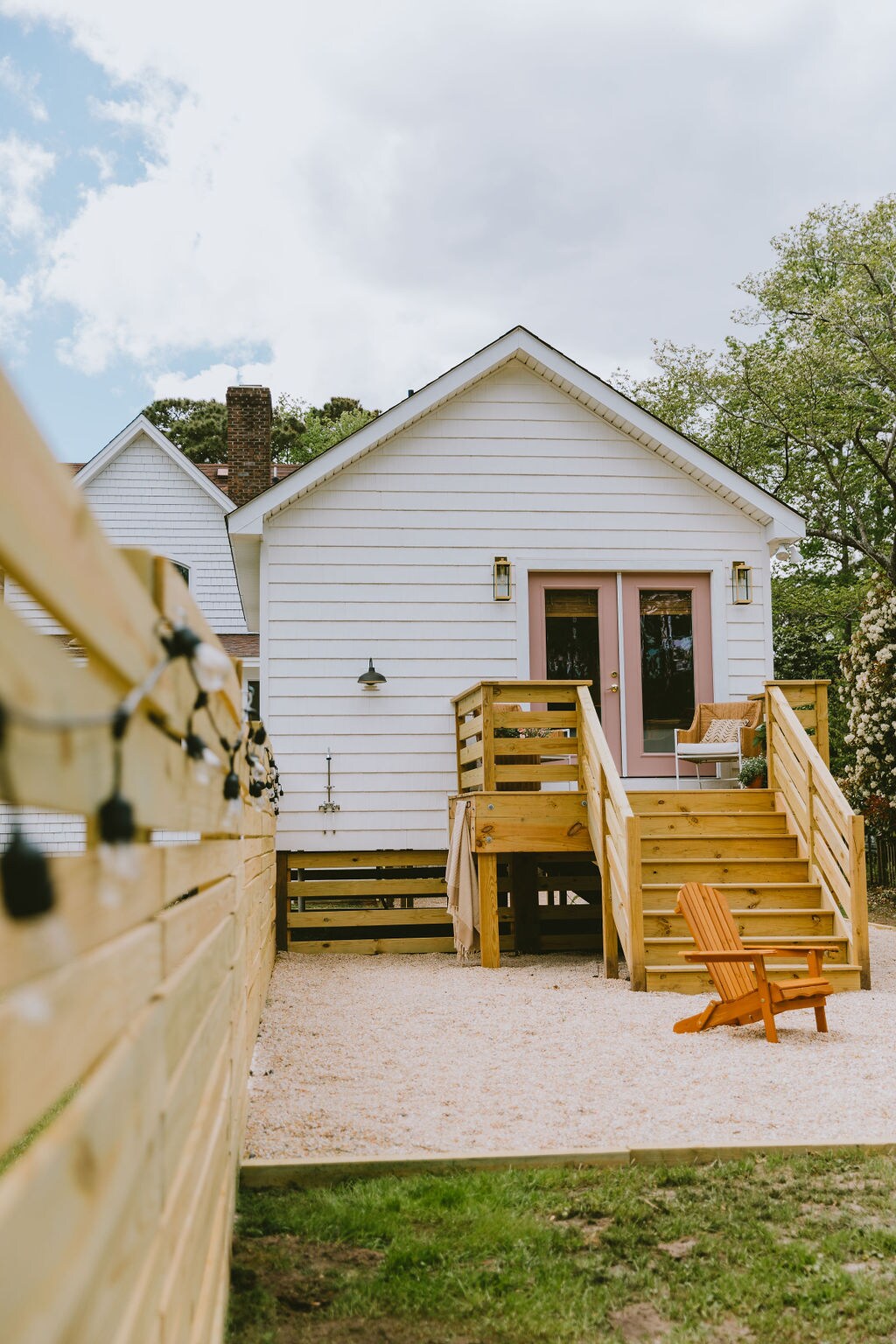 The exterior of the tiny house is shown, featuring a light-colored façade and a welcoming entrance with stairs. An Adirondack chair sits on the gravel patio, while string lights adorn the fence, creating a casual and inviting setting.