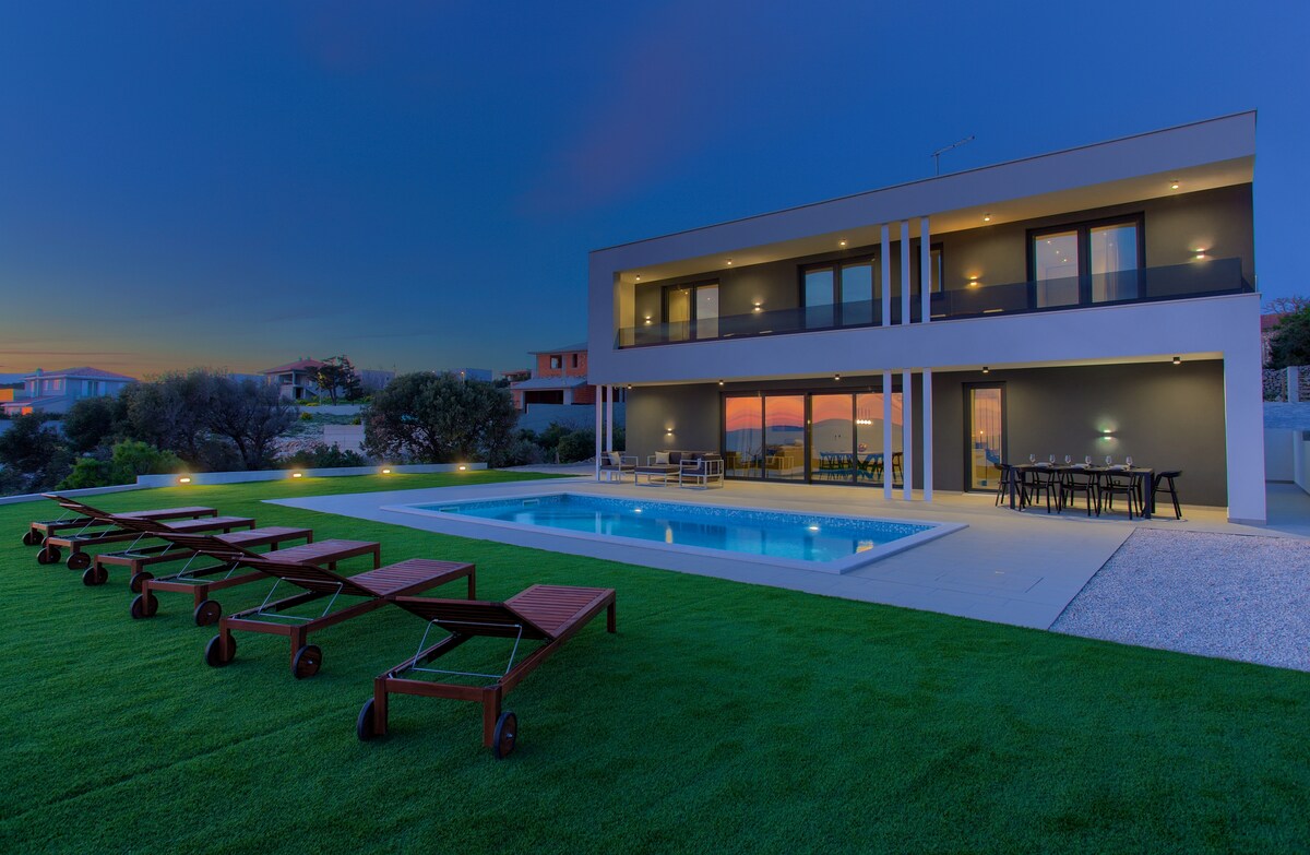 A modern villa is presented with an outdoor heated swimming pool surrounded by a well-manicured lawn. Several lounge chairs are positioned neatly nearby. The two-story structure features large windows that reflect the evening sky, creating a harmonious blend with the tranquil outdoor space.