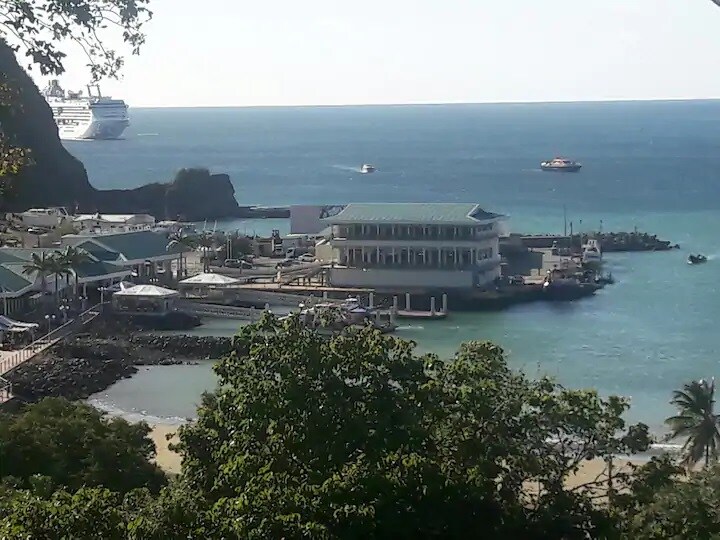 A scenic view of San Juan del Sur bay showcases calm waters and a distant cruise ship. The shoreline features a bustling marina with various boats and a prominent building along the water’s edge, surrounded by lush greenery and hills.