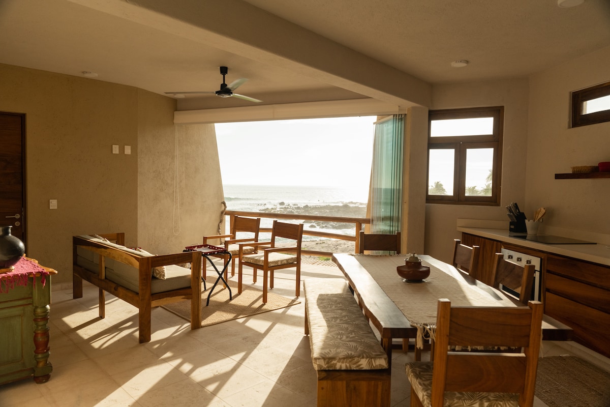 An open-concept living area features wooden furniture arranged for comfort, with large windows providing a view of the ocean. Natural light floods the space, and a ceiling fan is visible above. A dining table is set along one side, complemented by a kitchenette area.