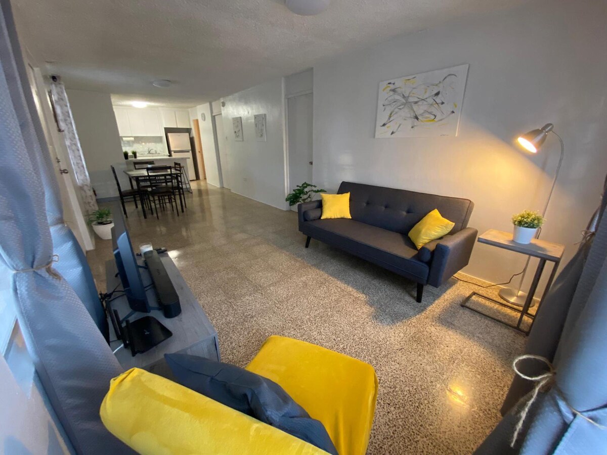 A living area features a gray sofa with yellow cushions and a small side table beside a floor lamp. A television is positioned nearby. The space is connected to a dining area visible in the background, with a table and chairs. Light streams through window treatments.