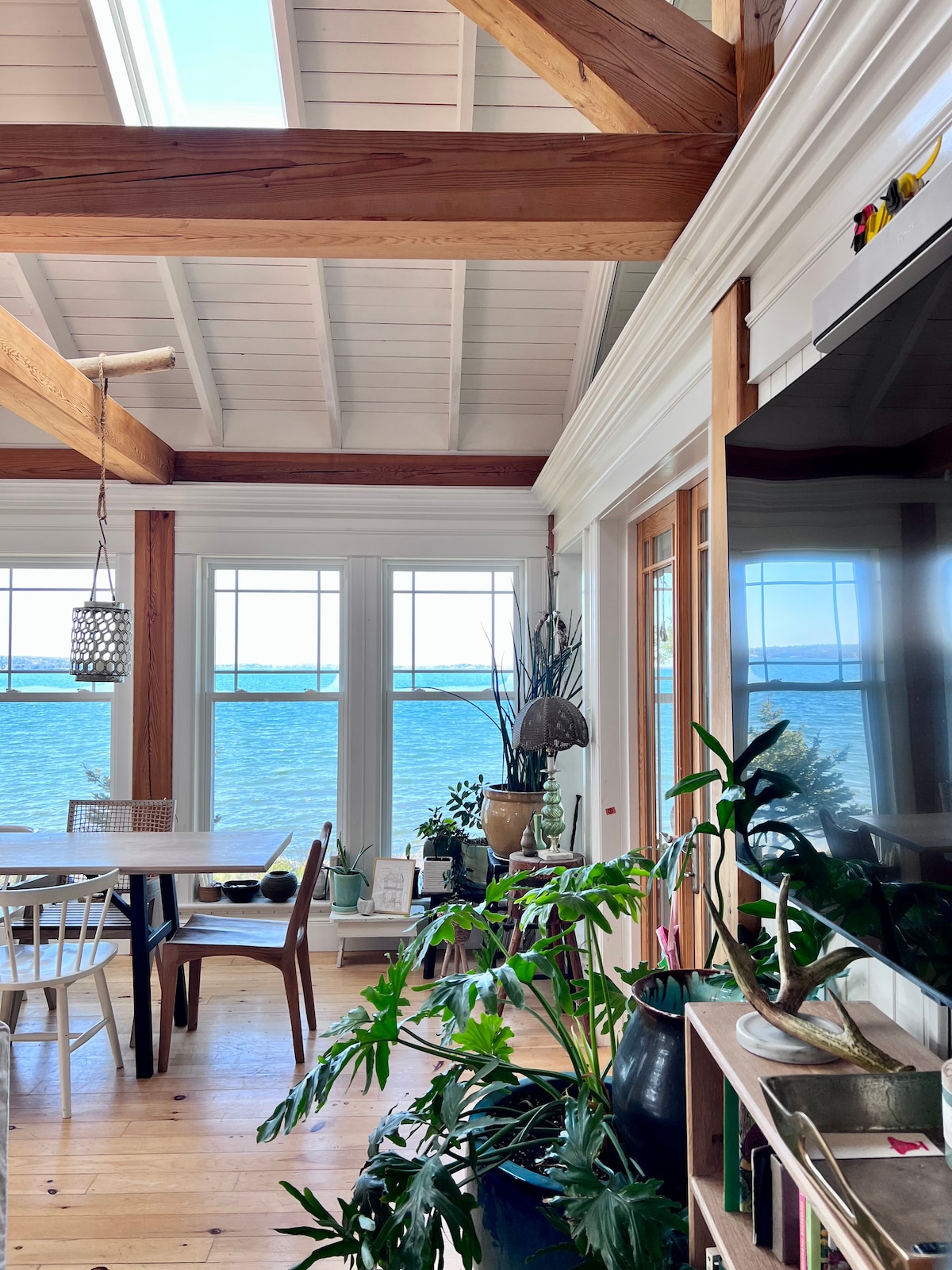 An airy open living area features a large dining table surrounded by chairs, with large windows overlooking the Atlantic Ocean. Various plants are positioned throughout the space, and natural light streams in, emphasizing the vaulted ceiling and wooden beams.