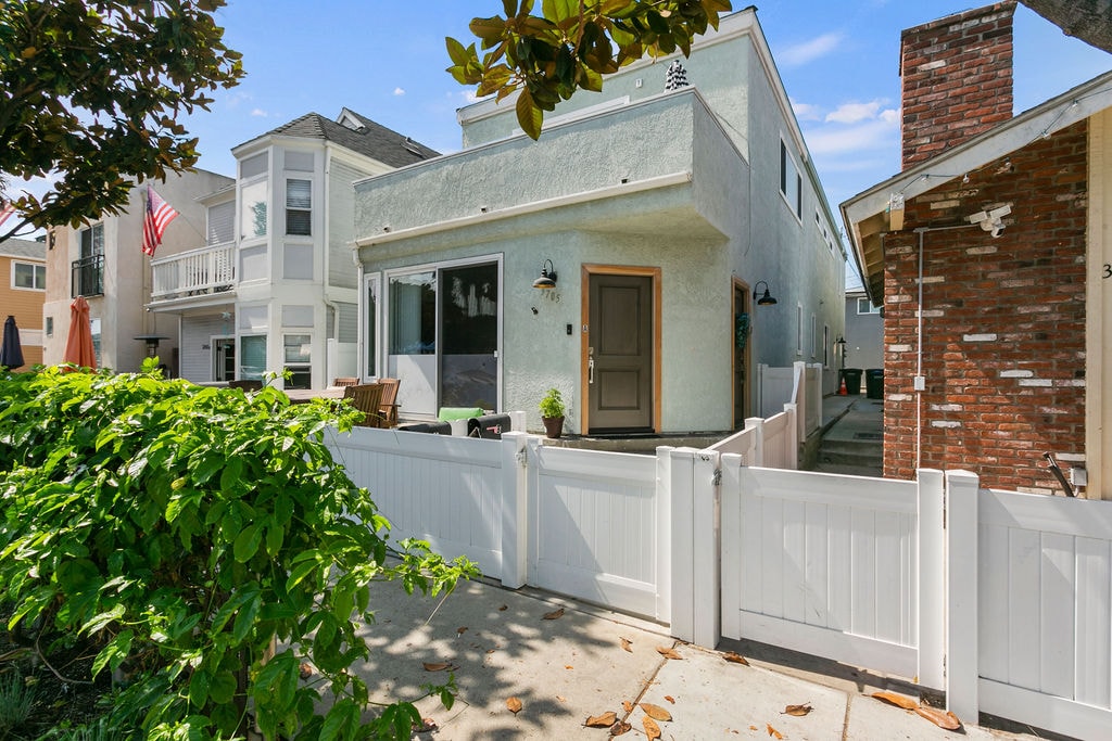 A two-story beach house is presented with a light-colored exterior, framed by a white picket fence. The entryway features a simple door and a welcoming porch area with seating. Lush greenery borders the pathway, enhancing the charm of the property.