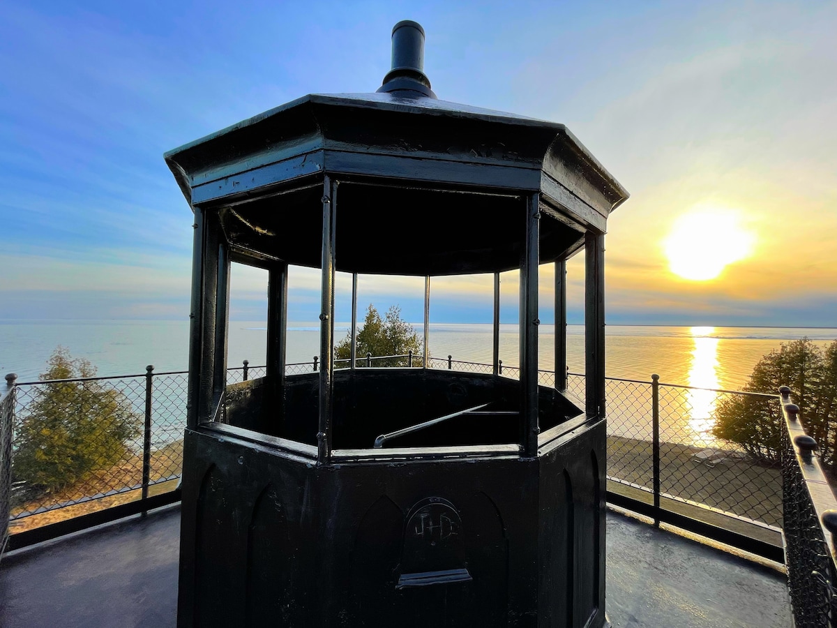 A black lantern structure is positioned atop a scenic overlook, providing expansive views of the lake and horizon. The warm hues of the sunset are reflected on the water's surface, creating a serene atmosphere. Foliage is visible in the foreground, adding to the natural setting.