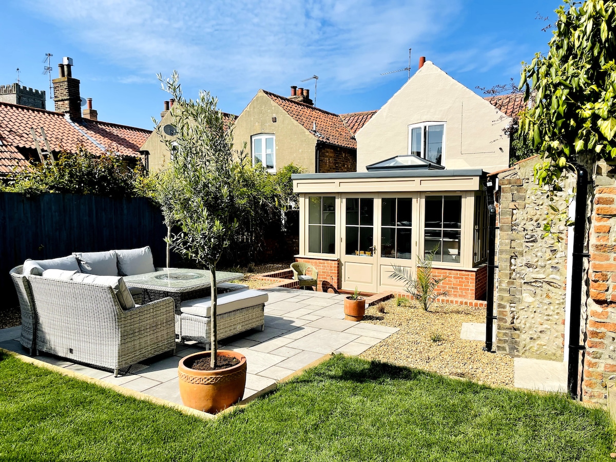 A sunny, enclosed garden features a spacious seating area adorned with a comfortable sofa set and a circular table. The garden path is framed by lush greenery, with a terracotta pot positioned at one corner, enhancing the outdoor relaxation space.