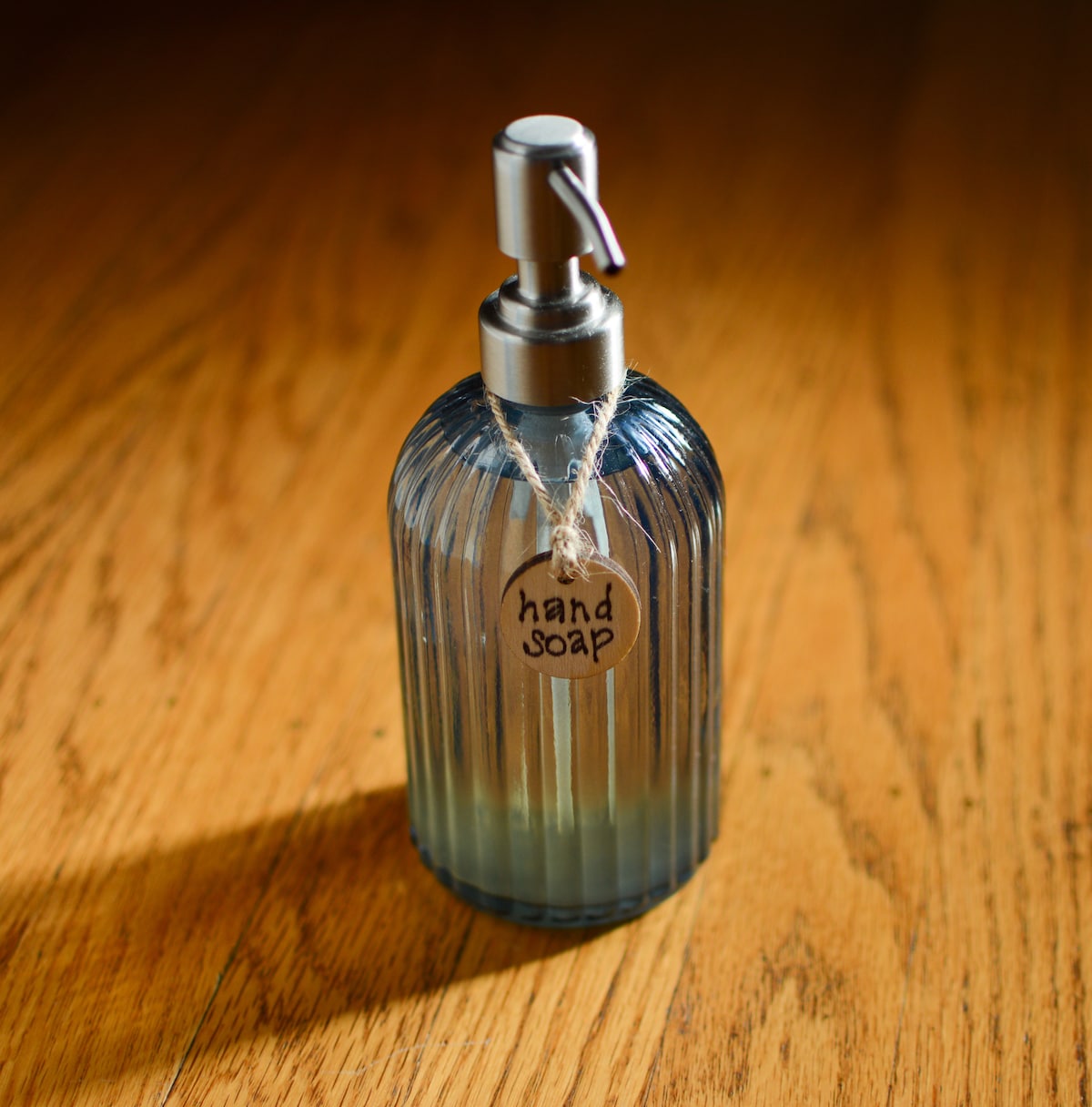 A glass hand soap dispenser with a ribbed design is placed on a wooden surface. The dispenser is partially filled, featuring a silver pump and a brown tag labeled 'hand soap.'