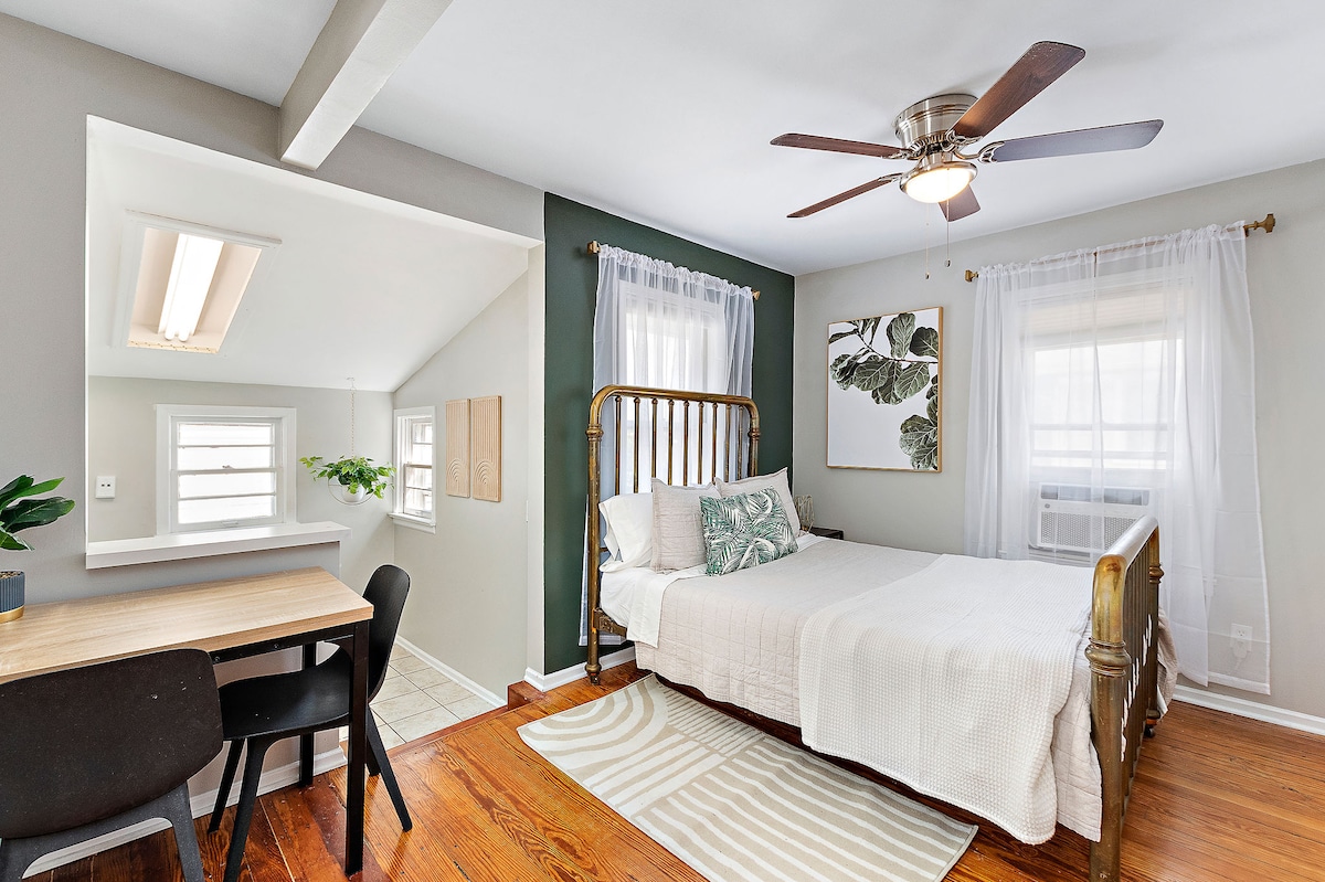 A studio space features a spacious bed with a light-colored cover, complemented by gray and green walls. A small dining area with a table and chairs sits nearby. Natural light filters through sheer curtains, enhancing the cozy feel of the room.