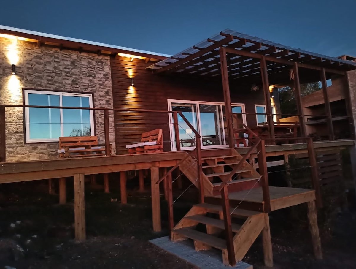 The exterior of the house is highlighted by a wooden deck featuring several seating areas. Soft lighting enhances the stone and wood facade, while steps lead down from the deck to the ground. Large windows reflect the evening sky, providing views of the surrounding nature.