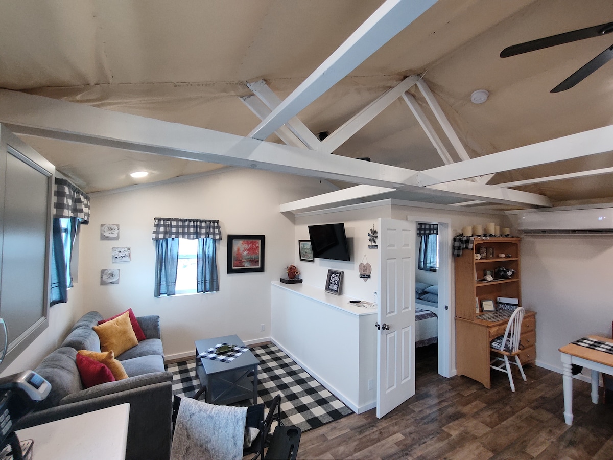 A spacious living area features a cozy gray sofa adorned with colorful pillows. A dark coffee table is centered on a checkerboard rug. A dining area with a wooden table and white chairs is visible, with a small workspace and a flat-screen TV mounted on the wall.