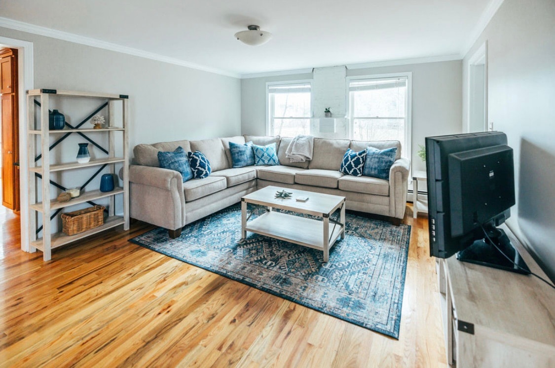 A spacious living area features a comfortable sectional sofa adorned with blue accent pillows, positioned near two windows that allow natural light to fill the room. A decorative area rug lies beneath a coffee table, while a television sits atop a light-colored media unit opposite the seating.