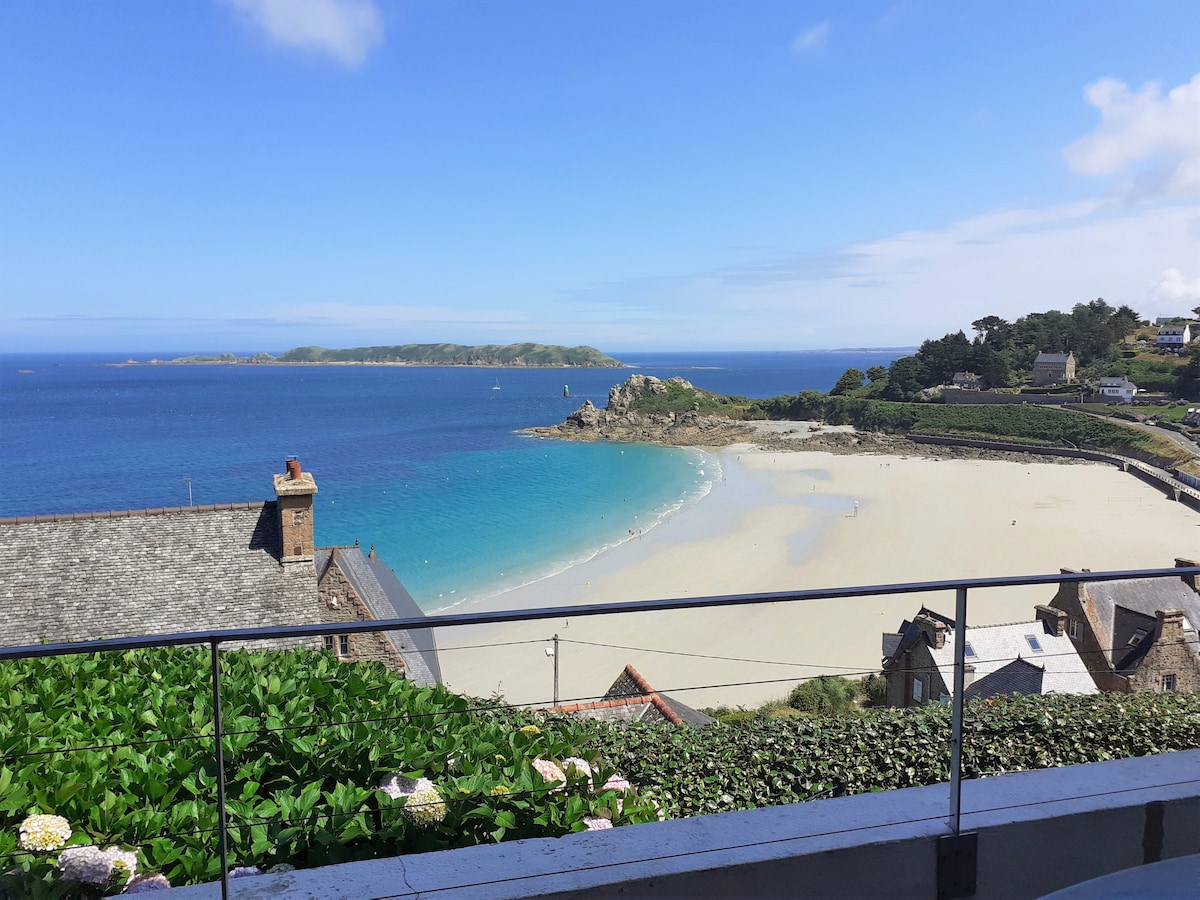 A panoramic view of the coastline is captured, featuring a sandy beach bordered by gentle waves. The foreground includes lush greenery along the terrace railing, while the expansive blue sky meets the ocean in the distance, highlighting the serene atmosphere of the area.