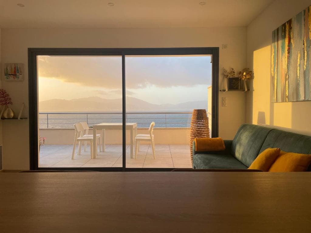 A spacious living area is visible, featuring a comfortable sofa with yellow cushions. Large glass doors open to a terrace with a dining table and chairs, offering views of the sea and surrounding mountains during sunset.