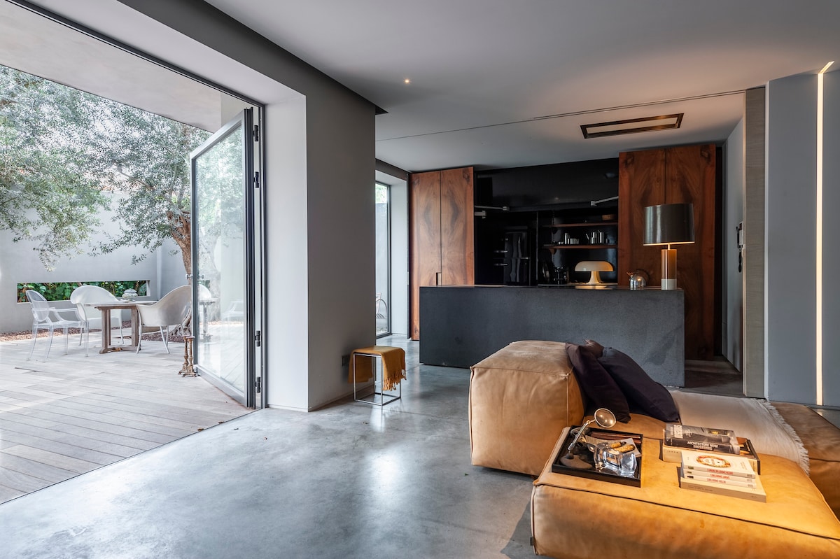 An open-plan living area featuring a cozy seating arrangement with a plush sofa and a coffee table. Large glass doors open to a wooden deck area, revealing a serene outdoor space with seating and greenery. A modern kitchen area is visible in the background.