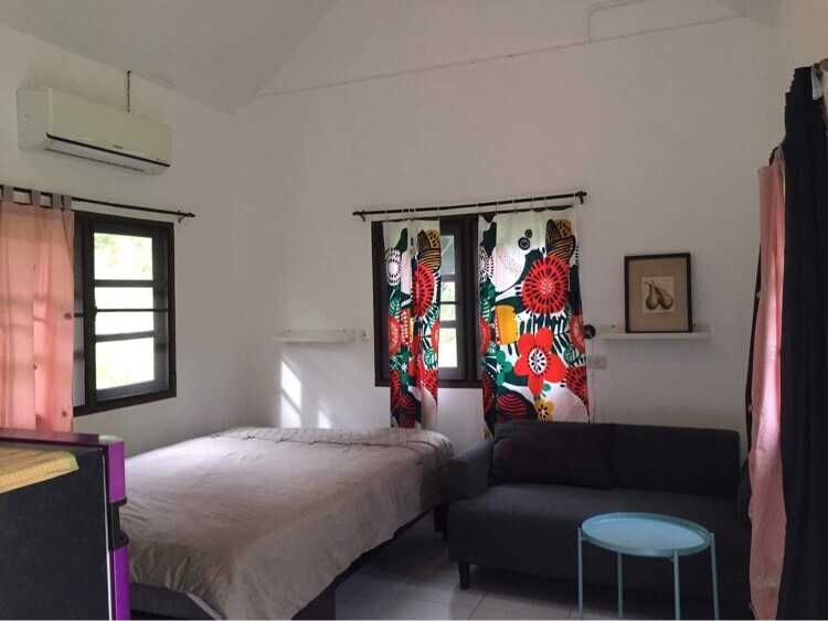 A bright room features a spacious bed, complemented by colorful floral curtains adorning two windows. A gray sofa adds comfort, while a light blue table provides a pop of color and functionality. The room is enhanced by an air conditioning unit mounted on the wall.