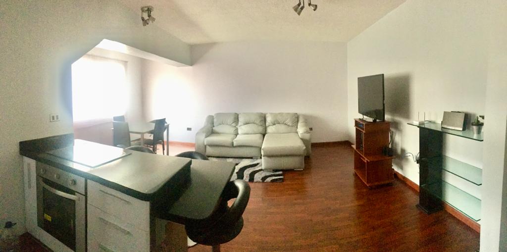 An open living space is featured with a comfortable sectional sofa positioned near a large window, providing natural light. A sleek television sits on a wooden stand, while a kitchen area with modern appliances is partially visible. A dining table is also present in the background.