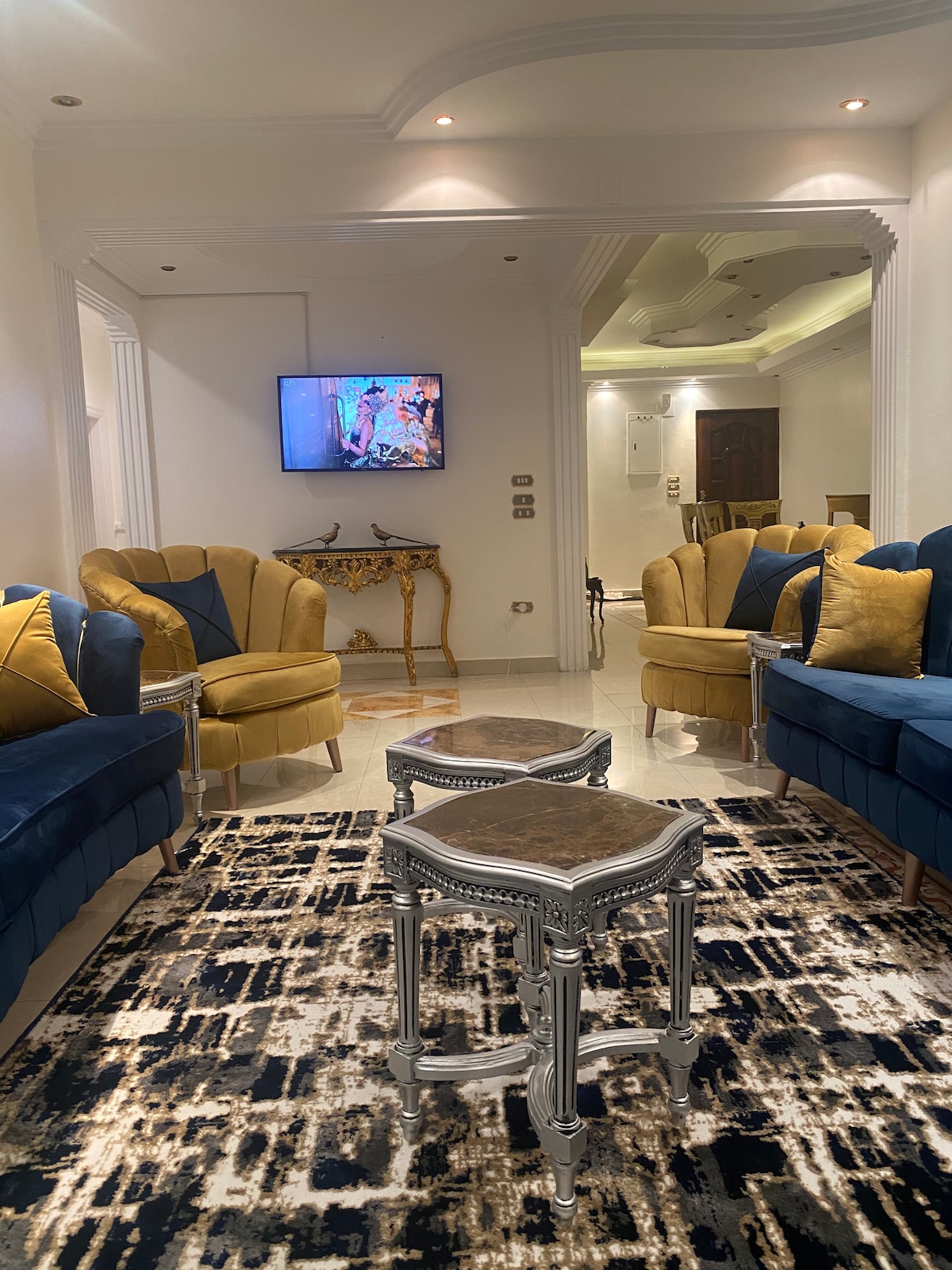 A living area is highlighted by a combination of yellow and blue armchairs arranged for conversation. A small ornate table sits in the center, with a textured rug covering the tiled floor. A television is mounted on the wall, visible to the seating area.