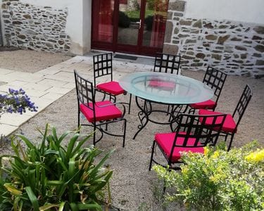 An outdoor dining area features a round glass table surrounded by six black chairs with vibrant red cushions. The setting is complemented by stone paving and lush greenery, providing a comfortable space for al fresco dining.