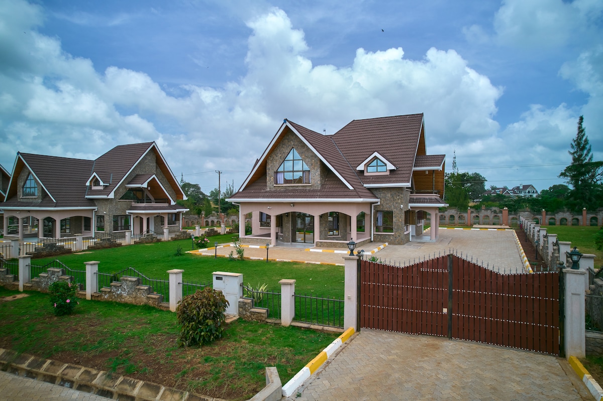 The exterior of a spacious villa is presented, featuring a gabled roof and stone accents. Surrounding landscaped gardens and a winding driveway lead to an inviting entrance, with sunlight filtering through fluffy clouds overhead.