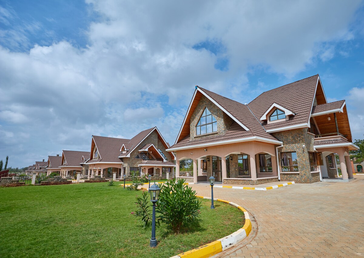 An elegant villa complex is showcased, featuring multiple structures with distinct gabled roofs and large windows. Lush green lawns surround the properties, while pathways paved with stone lead up to the entrances, blending seamlessly with the natural landscape.