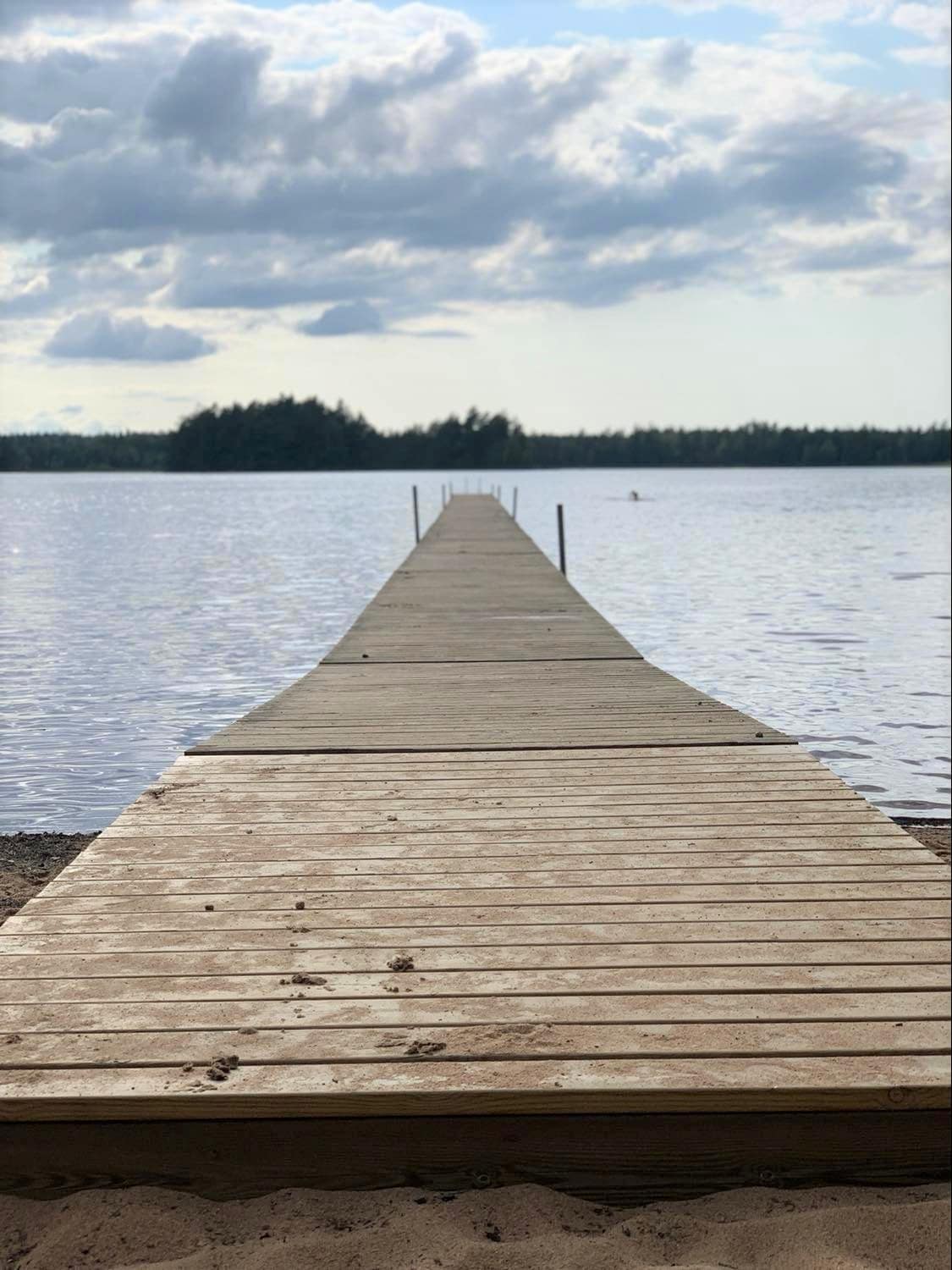A wooden dock extends into calm waters, leading towards a distant island framed by trees. The dock includes simple railings on either side and is set against a backdrop of a cloudy sky, creating a serene and peaceful environment.