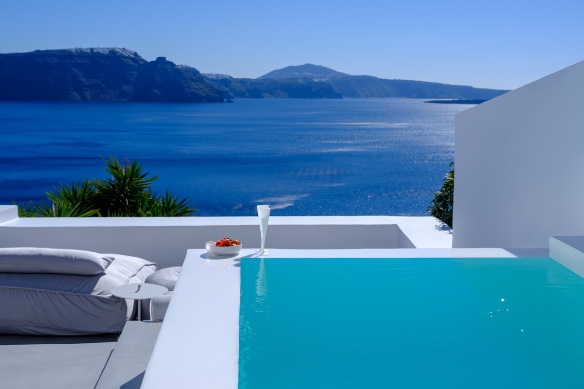 A serene outdoor space features a small plunge pool with clear turquoise water, overlooking a vast expanse of the Aegean Sea. A comfortable lounger is positioned nearby, along with a plate of fresh fruit and a glass, inviting relaxation amidst the stunning landscape.