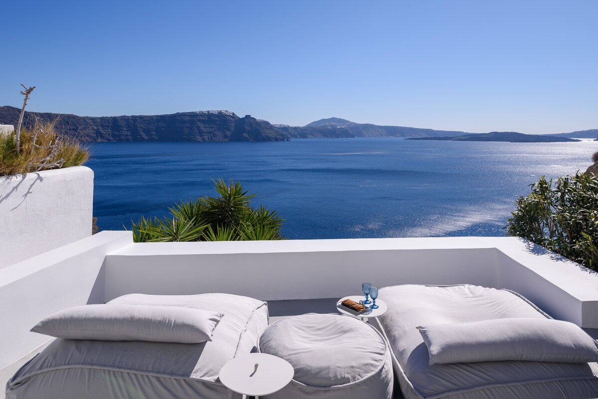 A serene seating area is presented on a private terrace, offering panoramic views of the deep blue Aegean Sea and surrounding cliffs. The cozy cushions invite relaxation, while a small side table holds refreshments, blending comfort with the stunning natural landscape.