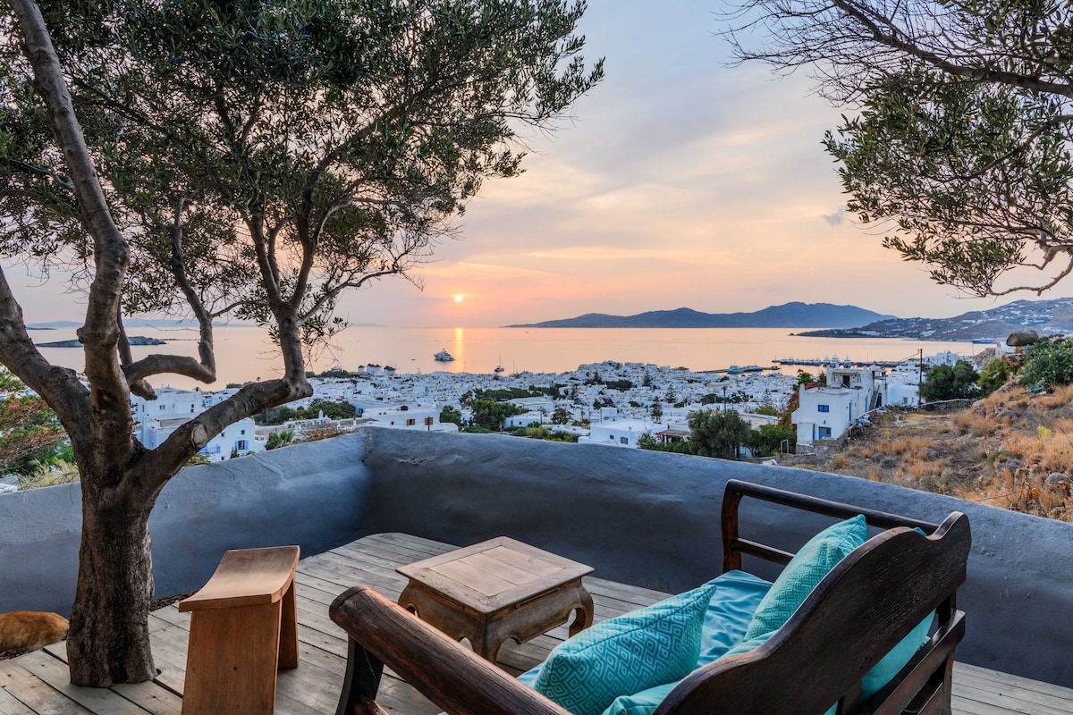 A wooden seating area with a turquoise cushion overlooks a serene landscape at sunset. The view includes distant islands and a calm sea, framed by olive trees. The deck features a small wooden table and showcases the natural colors of the evening sky.