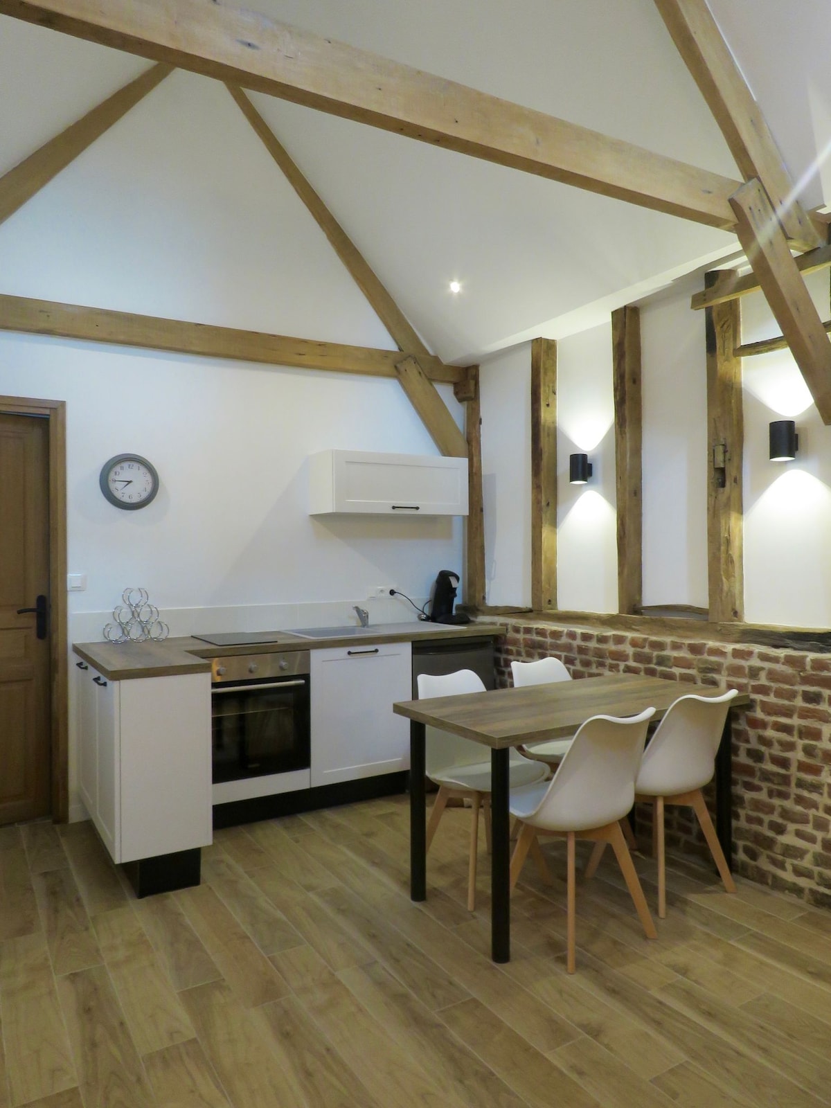 A bright kitchen area features modern appliances integrated into a minimalist design, complemented by natural wood beams overhead. A dining table is accompanied by four chairs, and a subtle brick accent wall enhances the space's character.