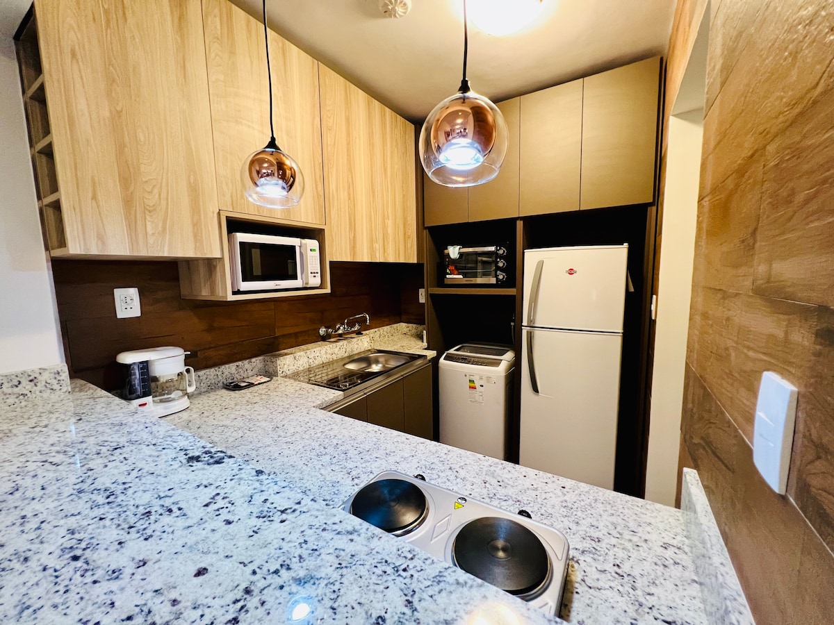 A modern kitchen features wooden cabinetry and sleek granite countertops. Two hanging light fixtures illuminate the space, highlighting a microwave, refrigerator, and stove. A coffee maker and various utensils are visible, suggesting a fully equipped cooking area.