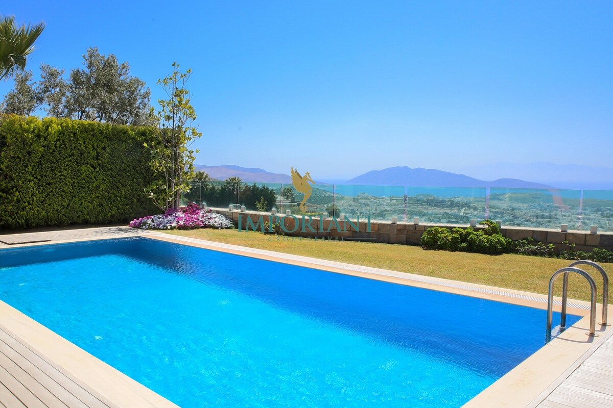 A well-maintained outdoor pool is surrounded by lush greenery and flowering plants. An expansive view of the ocean and distant mountains is visible in the background, providing a serene and inviting atmosphere for outdoor relaxation.