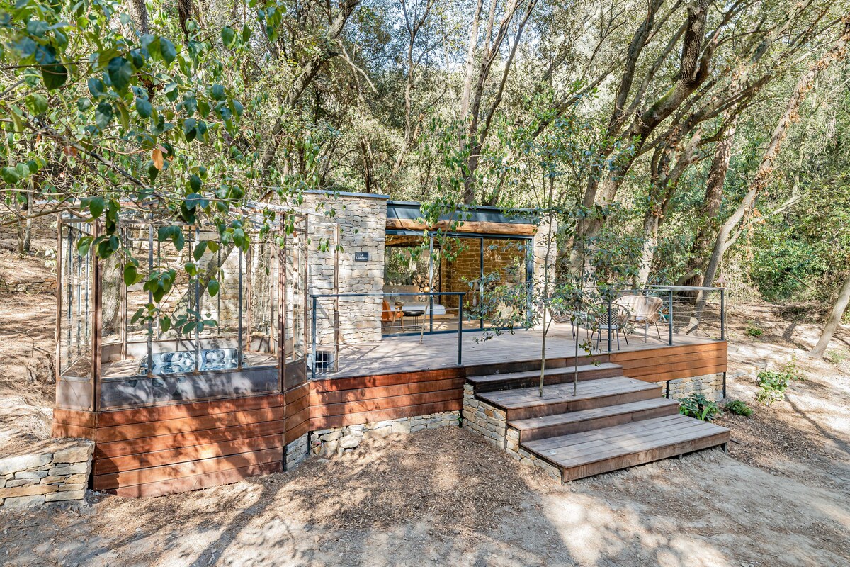 A spacious wooden terrace is surrounded by trees, providing a serene setting. Steps lead up to the terrace, which features a glass enclosure. The rustic stone structure behind blends harmoniously with the natural environment, offering a peaceful retreat.