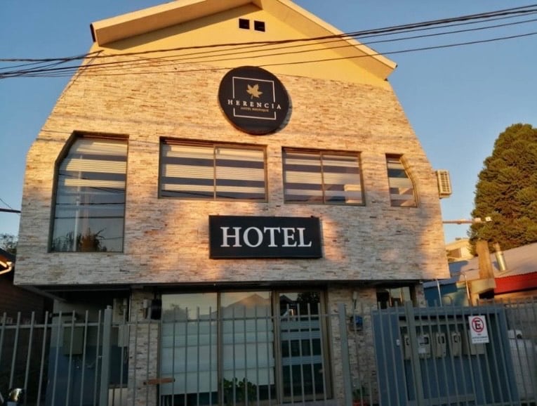 The exterior of a hotel is showcased with a modern design featuring a combination of stone and light-colored materials. Large windows allow natural light to enter, while a prominent sign displaying 'HOTEL' enhances visibility. A gated entrance is visible at the front.