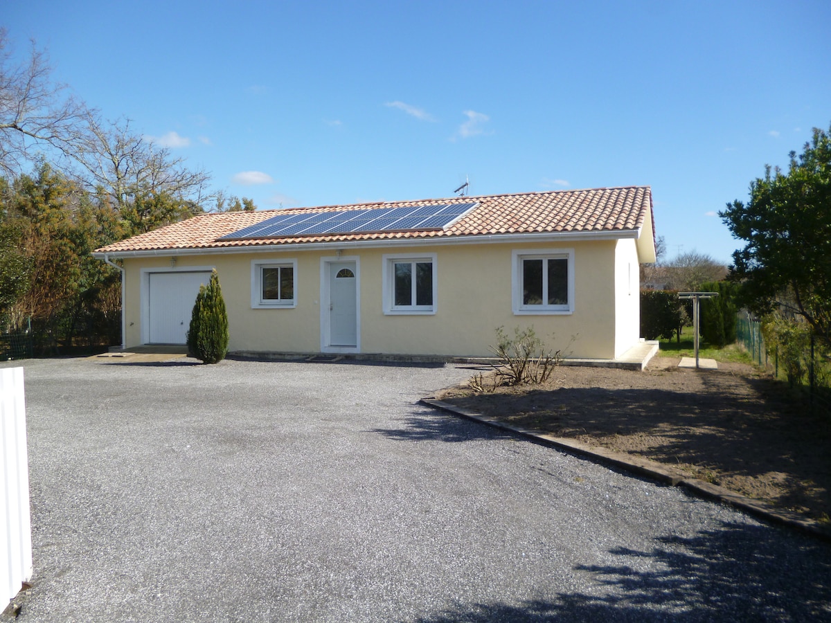 A single-story villa is presented, featuring a light-colored exterior and a sloped tiled roof. Solar panels are mounted on the roof. The front yard is gravelled, with a few bushes and landscaped areas visible, and a garage is attached to the left side of the house.