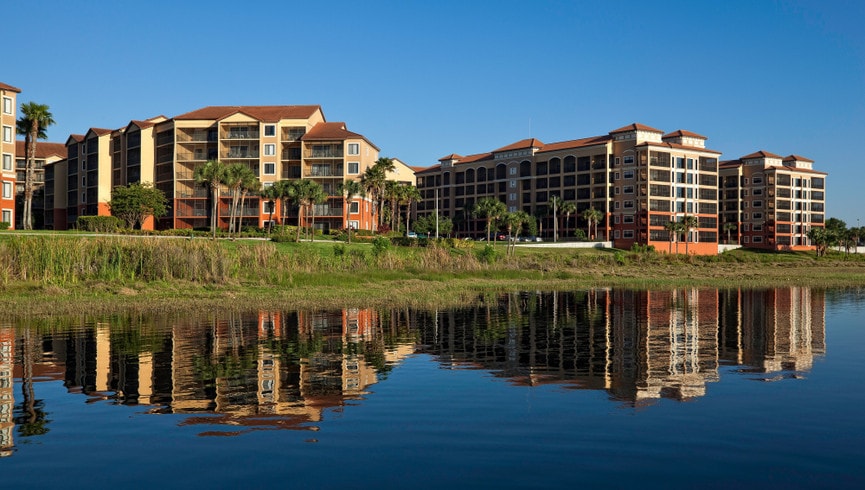 A modern resort complex is reflected in calm water, featuring multiple buildings with balconies and lush landscaping. Clear skies and palm trees enhance the serene environment, creating a tranquil setting for relaxation.