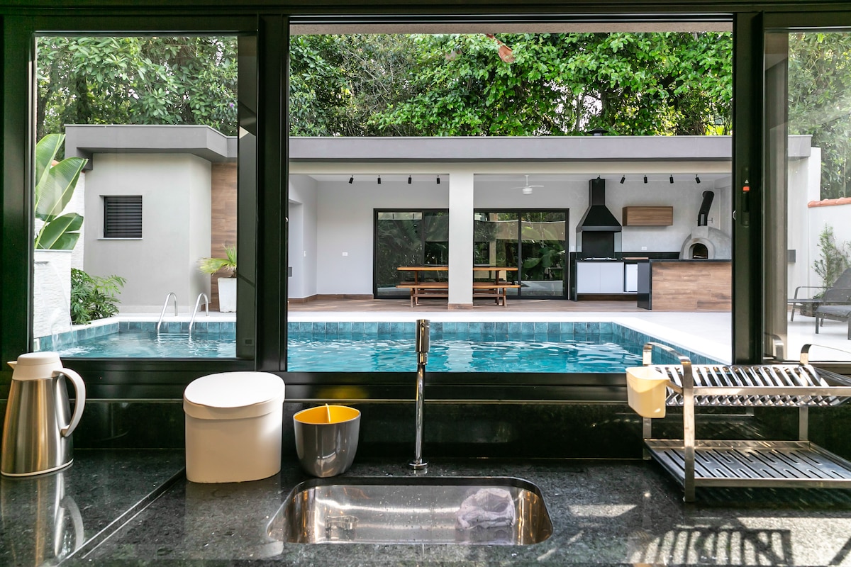 The kitchen area is visible, featuring dark countertops and a sleek sink with modern fixtures. Large windows provide a view of the outdoor space, which includes a heated infinity pool, seating area, and barbecue. Natural greenery surrounds the scene, enhancing the connection with nature.